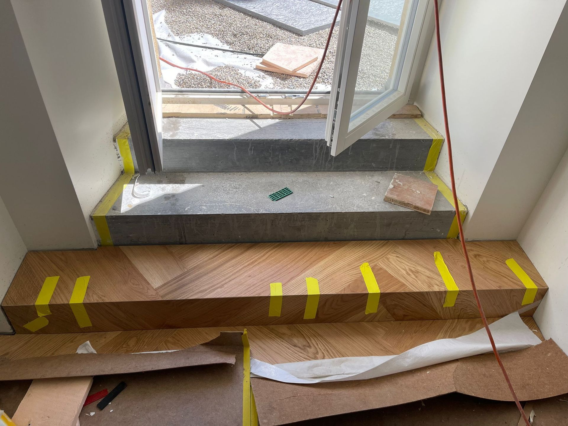 Une zone de construction montrant un seuil en pierre menant à une porte avec un escalier en bois nouvellement installé en dessous, balisé avec du ruban adhésif pour les travaux.