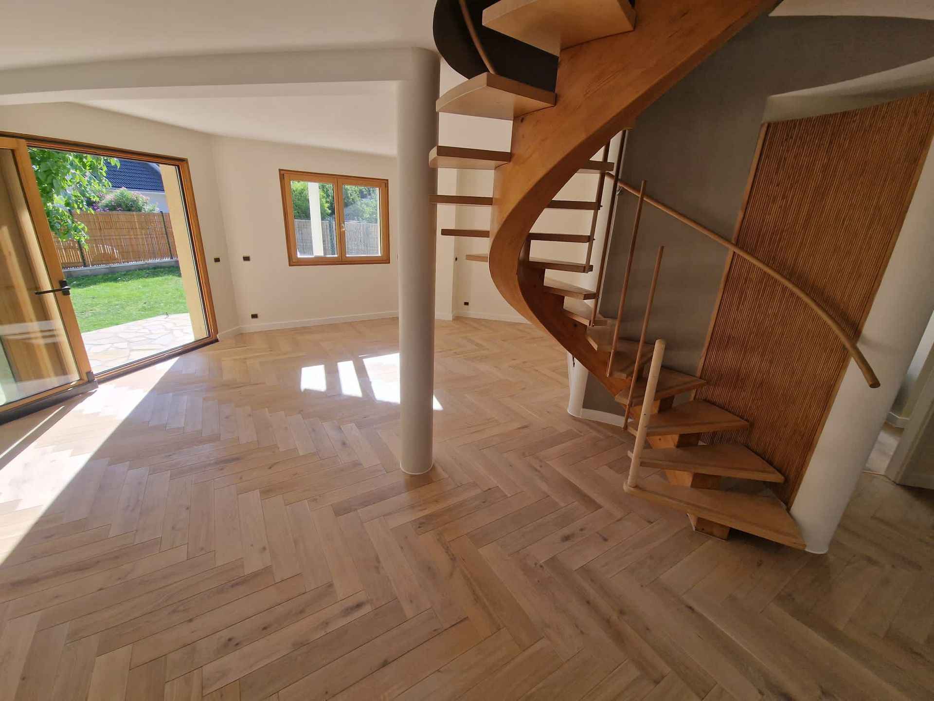 Une pièce spacieuse avec un parquet en chevrons, un escalier circulaire, une colonne de soutien et une porte donnant sur une cour.