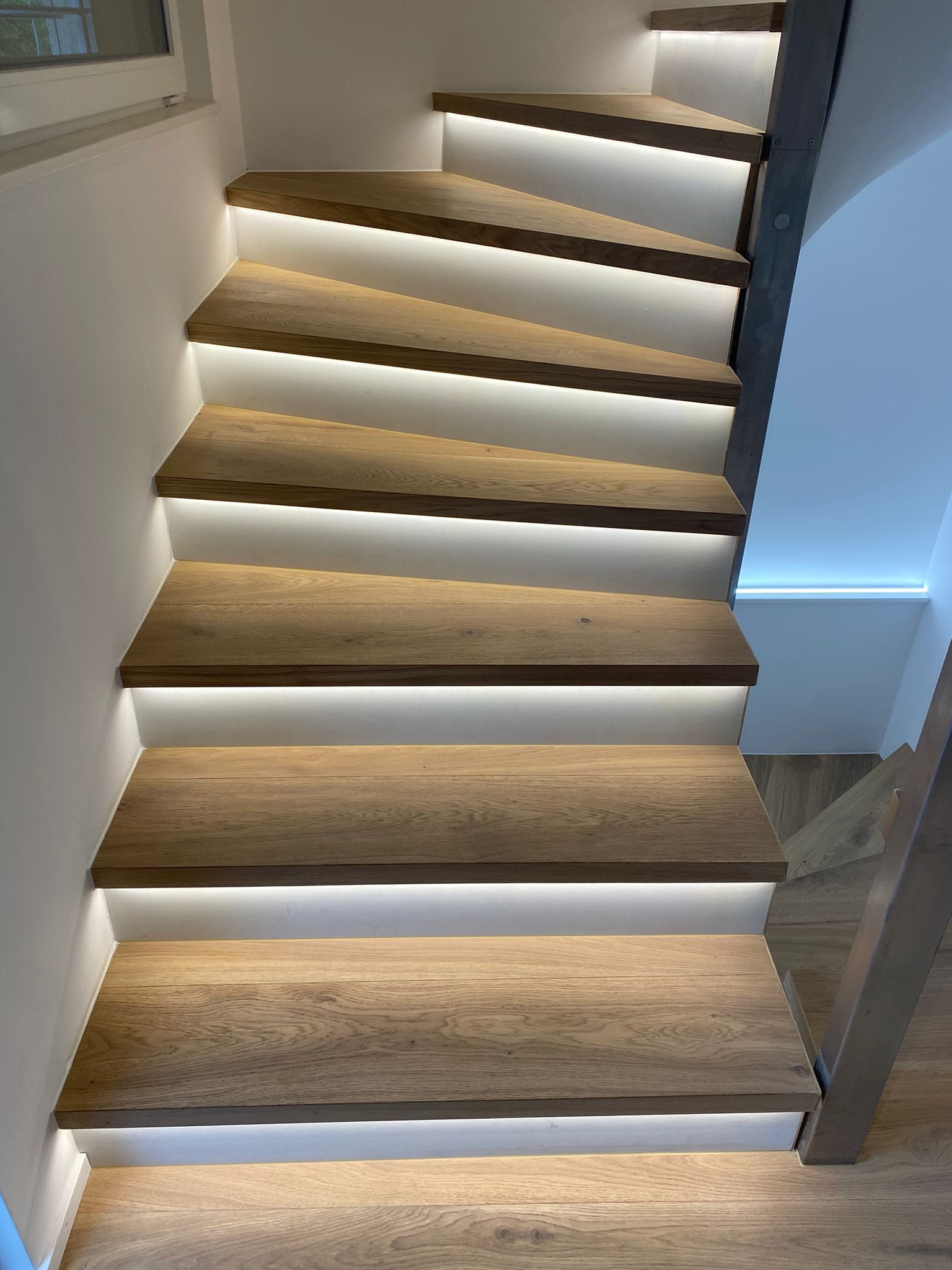 Un escalier avec des marches en bois clair, des contremarches blanches et un éclairage LED sous les marches.