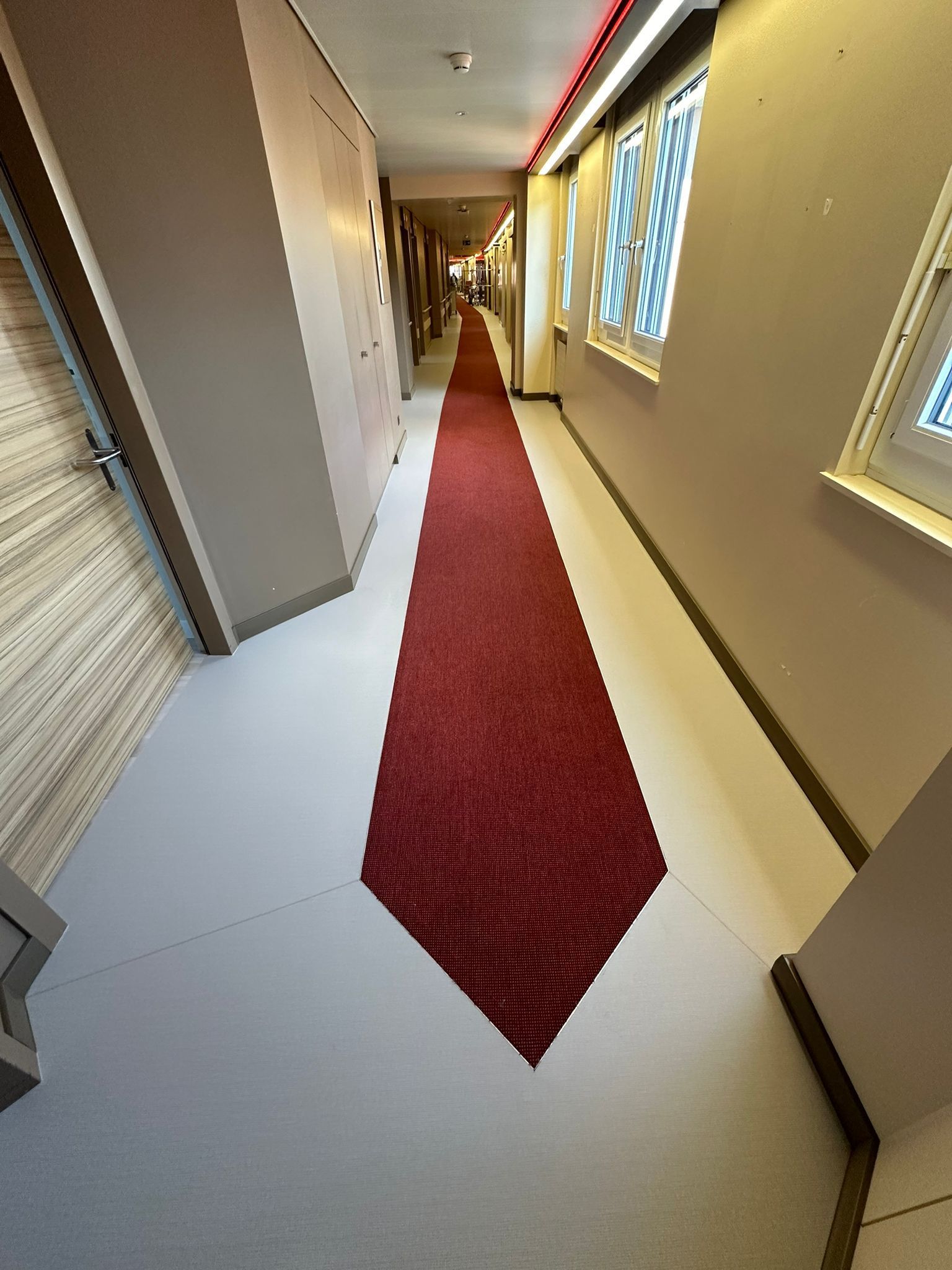 Un couloir étroit au sol clair et un long tapis rouge effilé menant à une porte au loin.