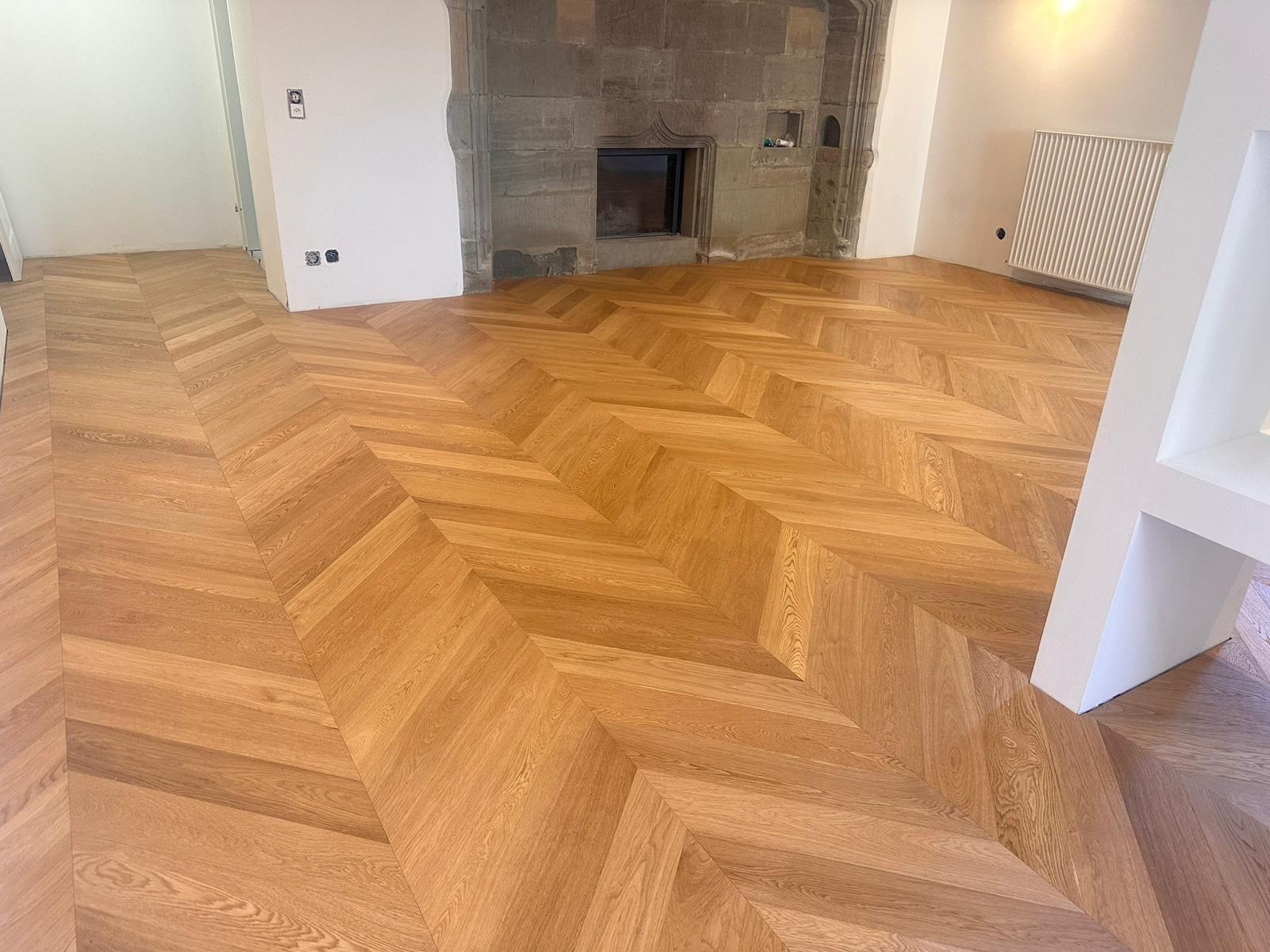 Un parquet clair à chevrons habille une pièce intérieure avec une cheminée en pierre en arrière-plan.