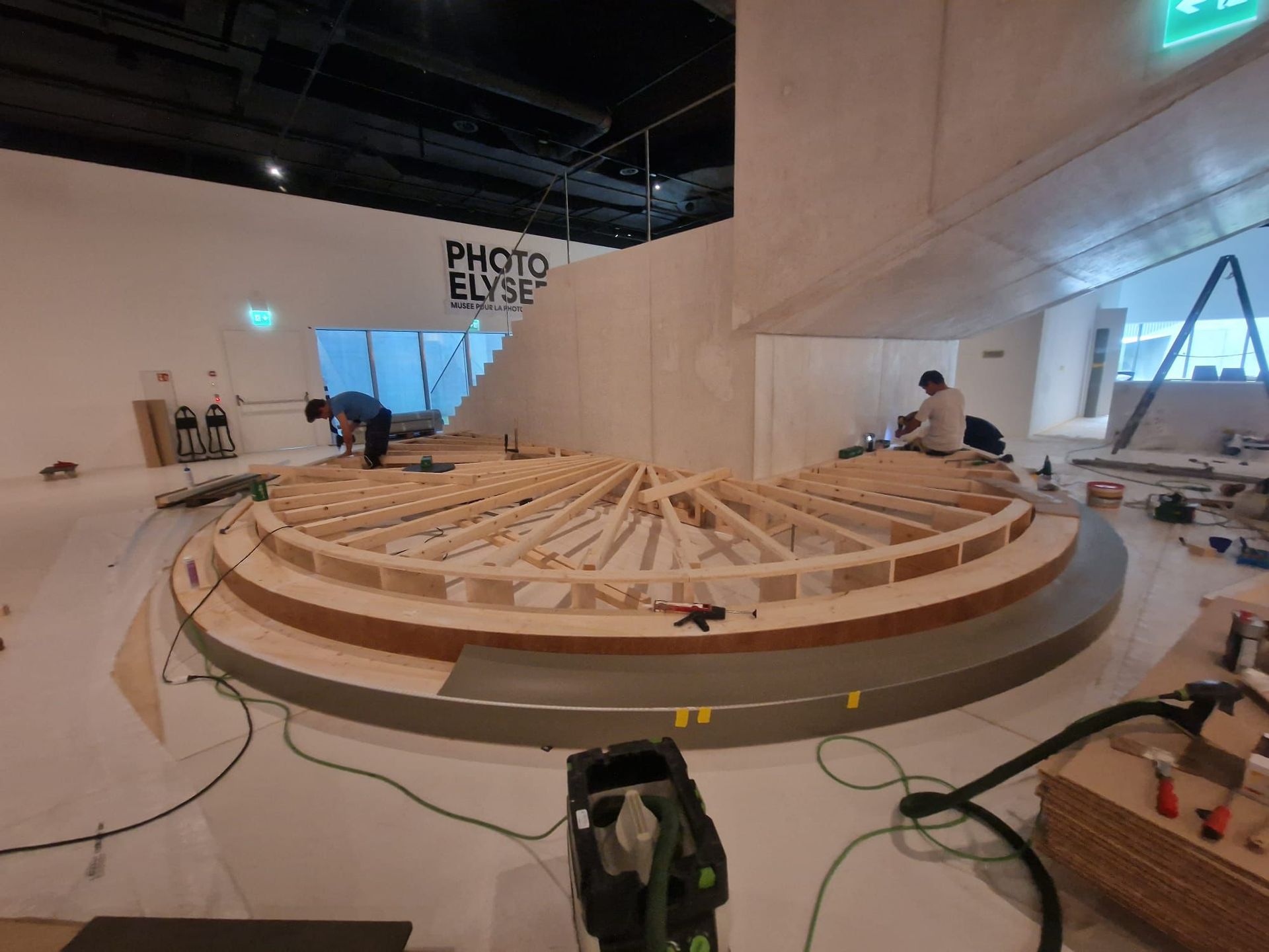 Deux ouvriers construisent une grande plateforme en bois incurvée à l'intérieur d'une galerie moderne où l'on peut lire « PHOTO ELYSEE » sur le mur.