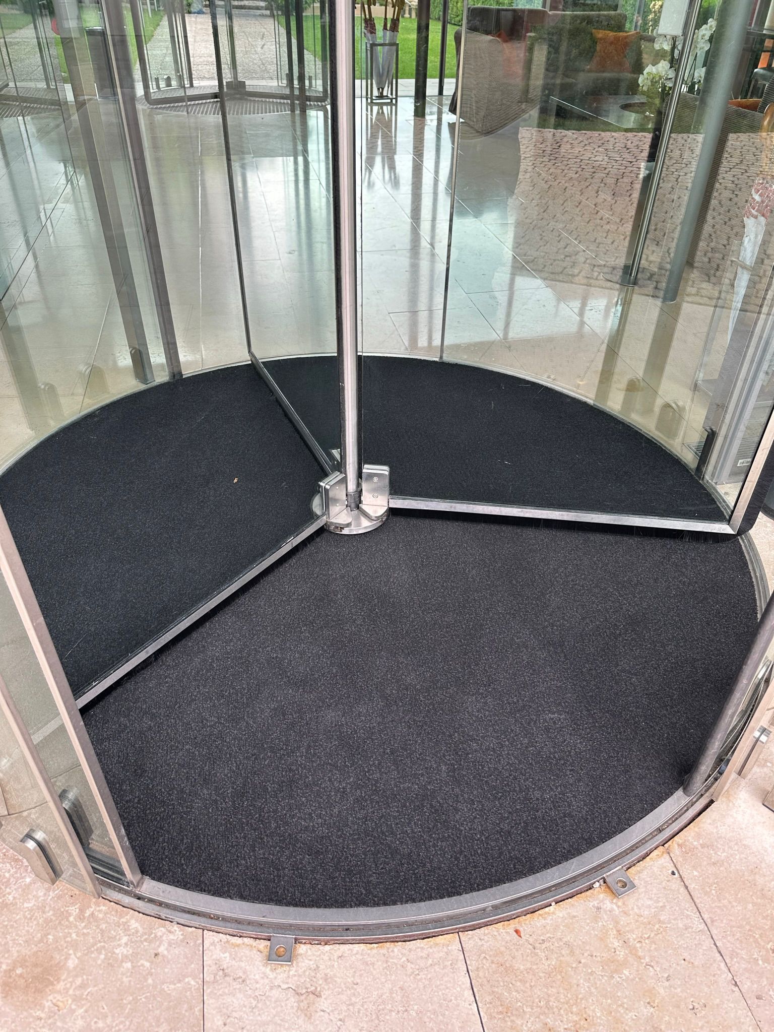 Une entrée à porte tournante avec un sol en tapis noir, des parois en verre et un poteau métallique central.