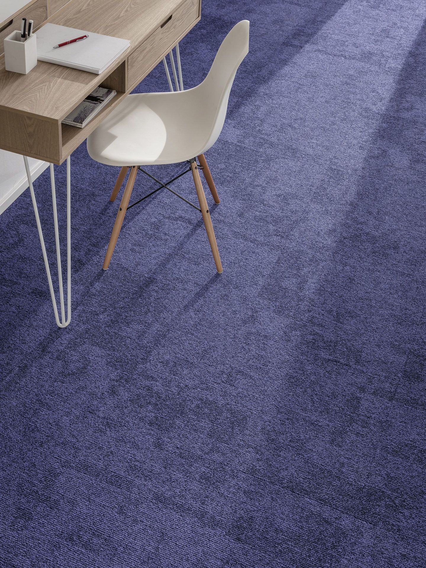 Une chaise blanche et un bureau en bois clair avec de la papeterie sont posés sur une moquette texturée violet foncé.