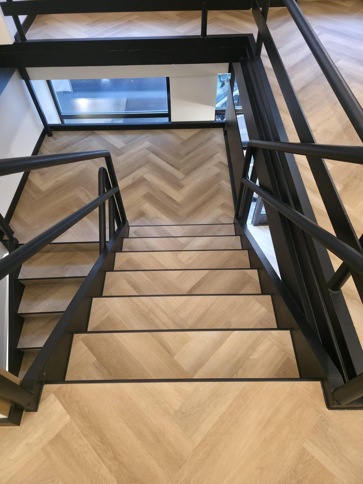 Vue en plongée d'un escalier en bois avec parquet à chevrons et rampes en métal noir.