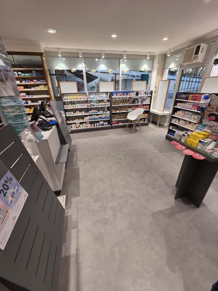 Vue intérieure d'une pharmacie lumineuse présentant des étagères de produits de santé, un comptoir central et un sol gris clair.