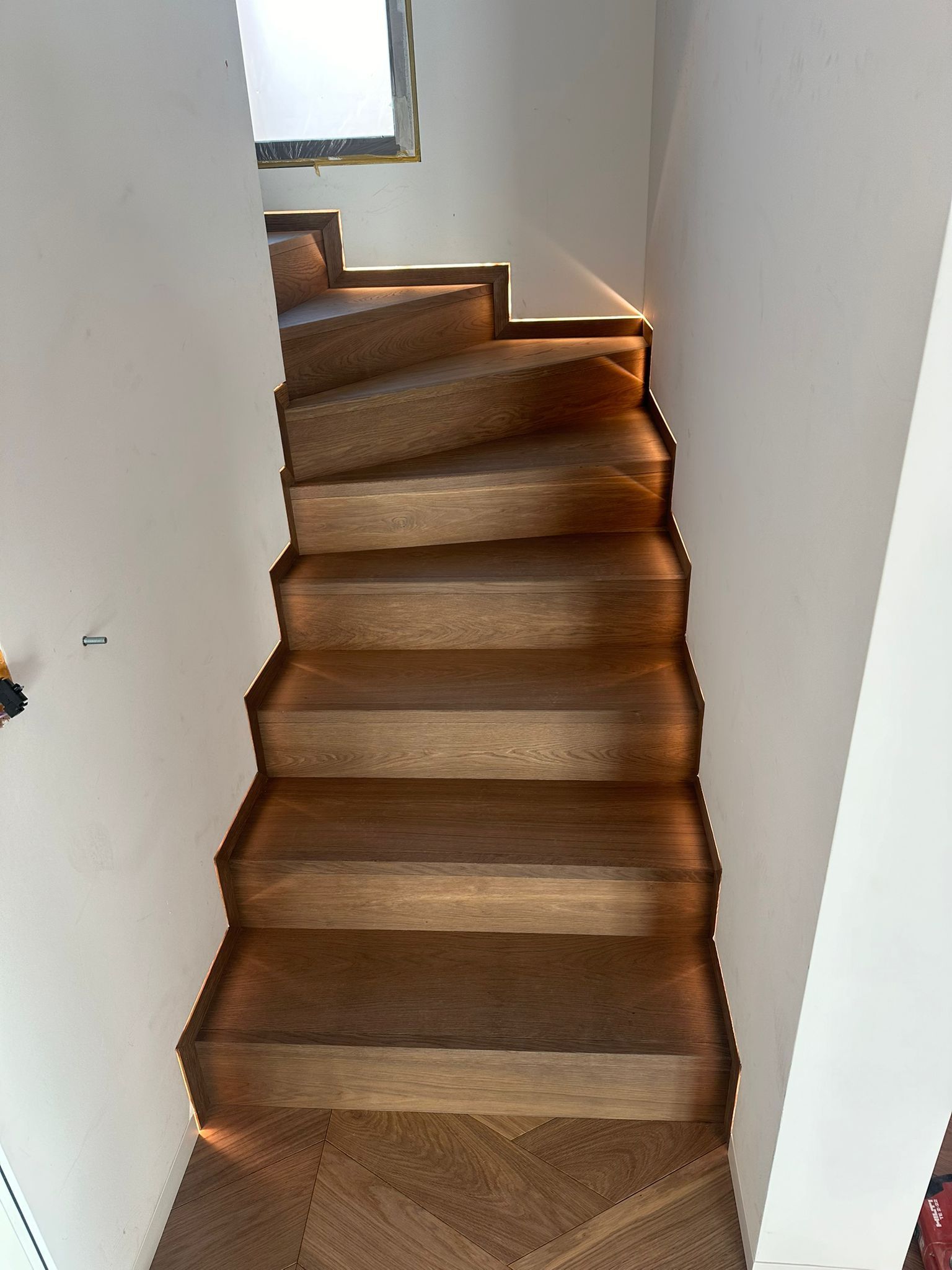 Un escalier en bois avec éclairage d'ambiance chaleureux intégré sous chaque marche, situé dans un couloir étroit aux murs blancs.