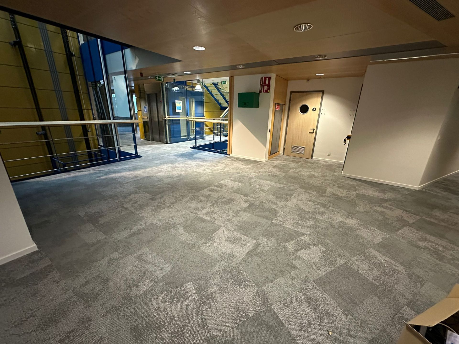 Un espace de bureaux intérieur lumineux et moderne, avec une moquette grise à motifs, un ascenseur en verre et une porte fermée au fond.
