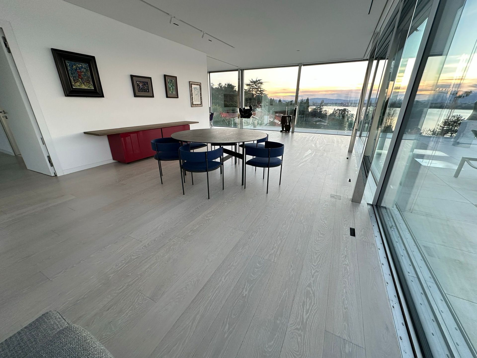 Salle à manger minimaliste avec chaises bleu foncé, table ronde en bois, buffet rouge et œuvre d'art murale donnant sur un lac.