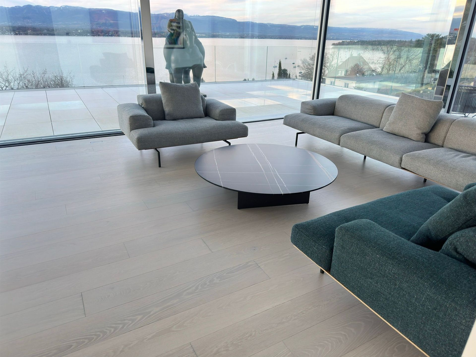 Un salon moderne avec des canapés en tissu gris et turquoise, une table basse ronde et une grande baie vitrée offrant une vue imprenable.