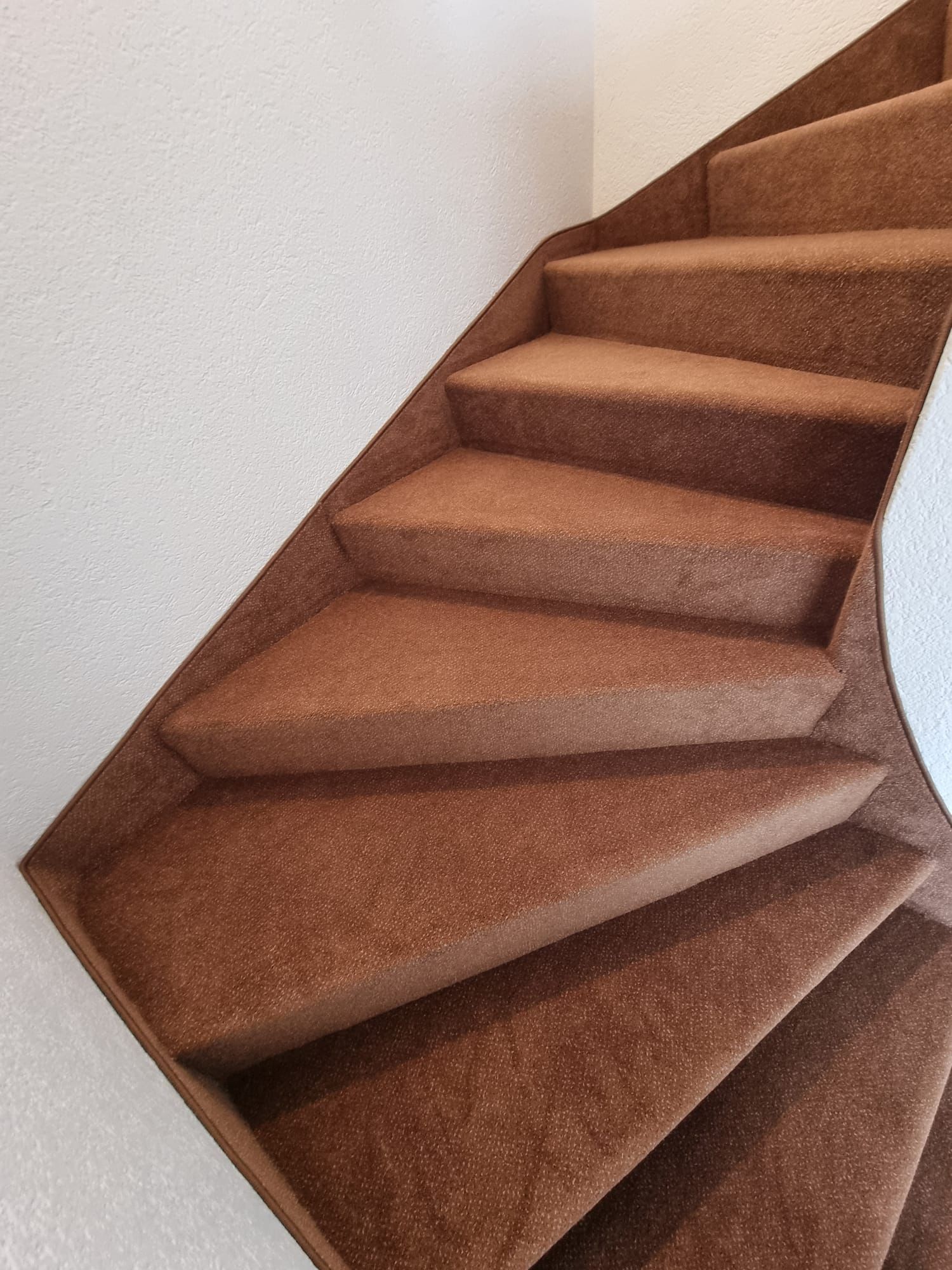 Un escalier aux marches en moquette texturée marron, incurvées vers le haut contre un mur blanc.