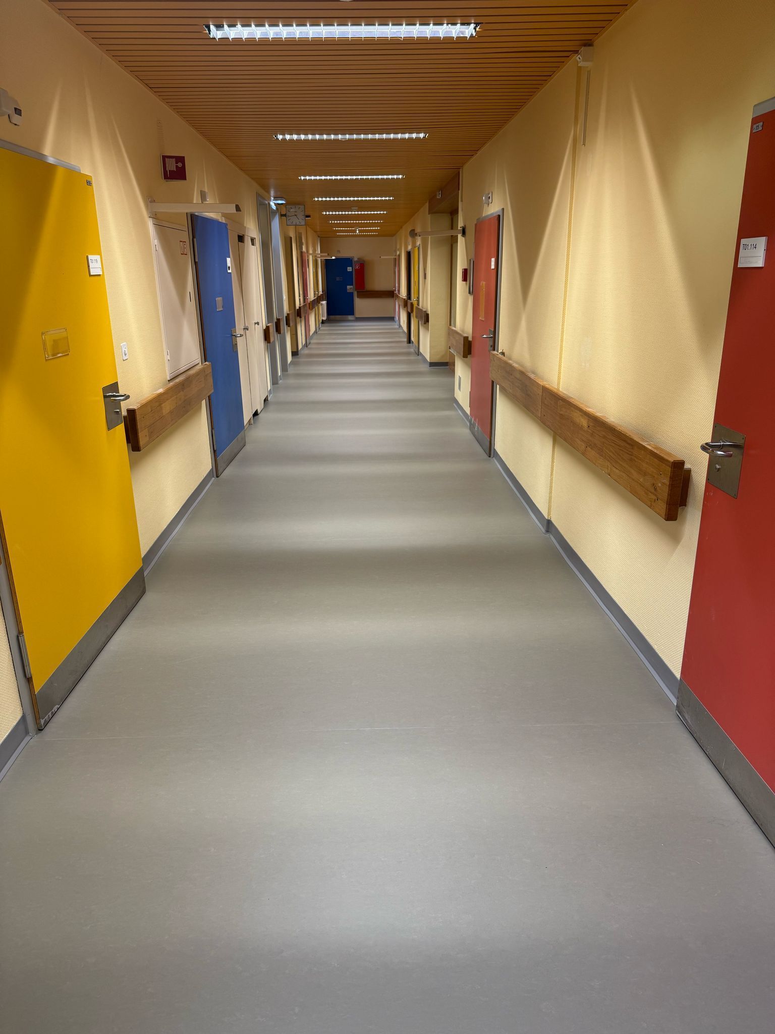 Un long couloir d'hôpital étroit, aux murs jaune clair, au sol gris et aux portes aux couleurs vives : rouge, jaune et bleu.