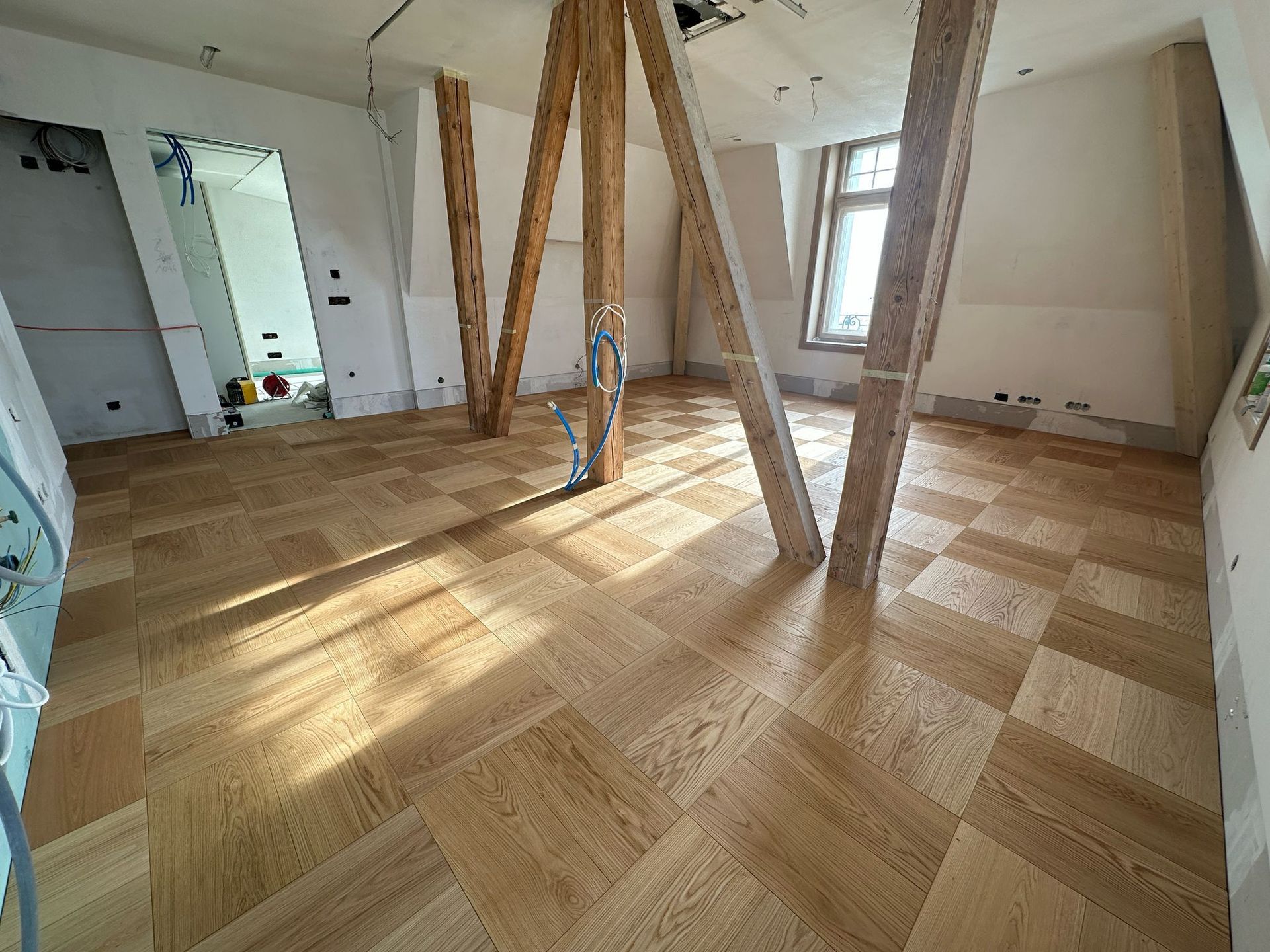 Une pièce intérieure en construction avec un parquet clair à carreaux, des poutres de soutien en bois et des murs blancs.
