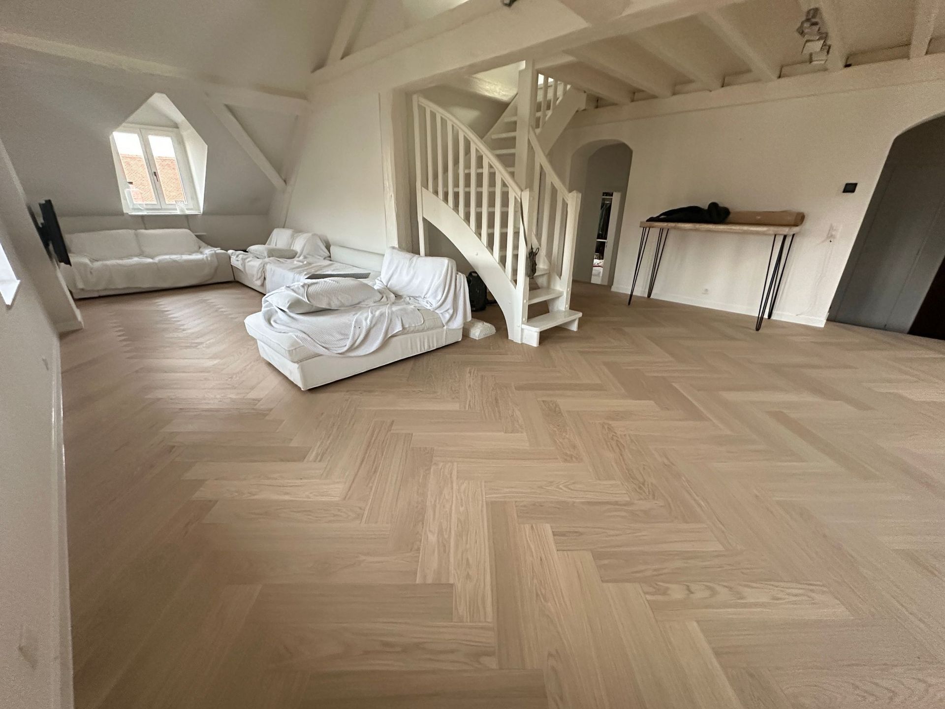 Un salon mansardé lumineux et spacieux avec parquet clair à chevrons, canapés blancs et escalier en bois blanc.