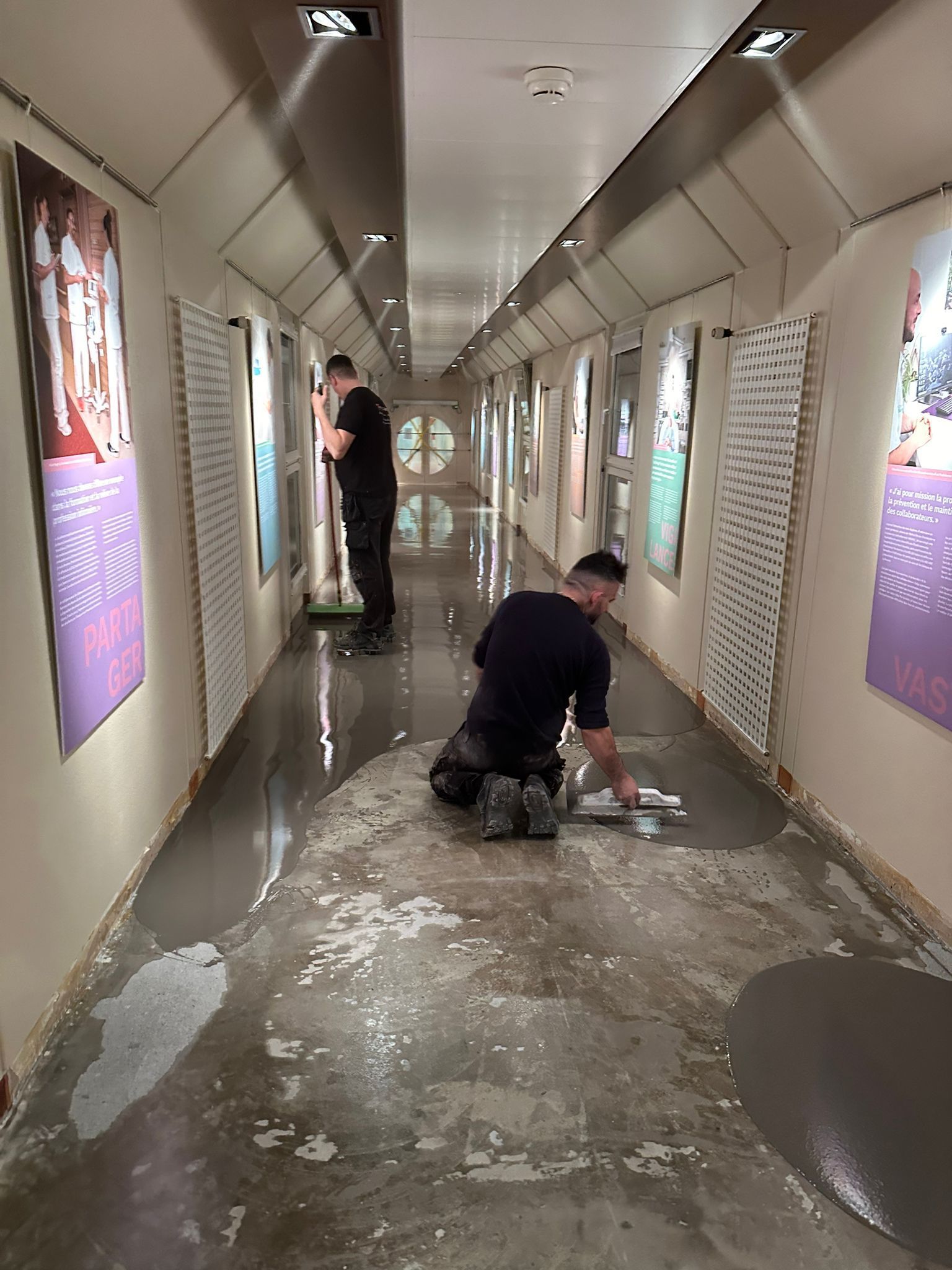 Deux personnes appliquent un enduit de nivellement gris et lisse dans un couloir étroit et lumineux, tapissé d'affiches murales.
