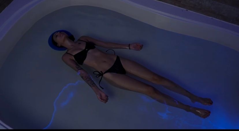 Person floating in a dim blue-lit tub, wearing a black bikini and snorkel mask.