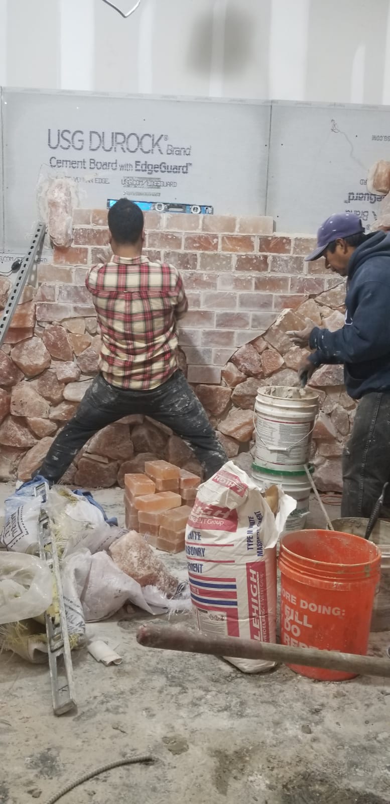 Two workers plastering a brick wall indoors, with buckets, cement bags, and debris on the floor.