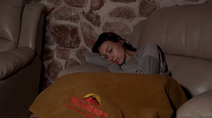 Person asleep on a couch beside a stone wall, covered with a brown blanket.