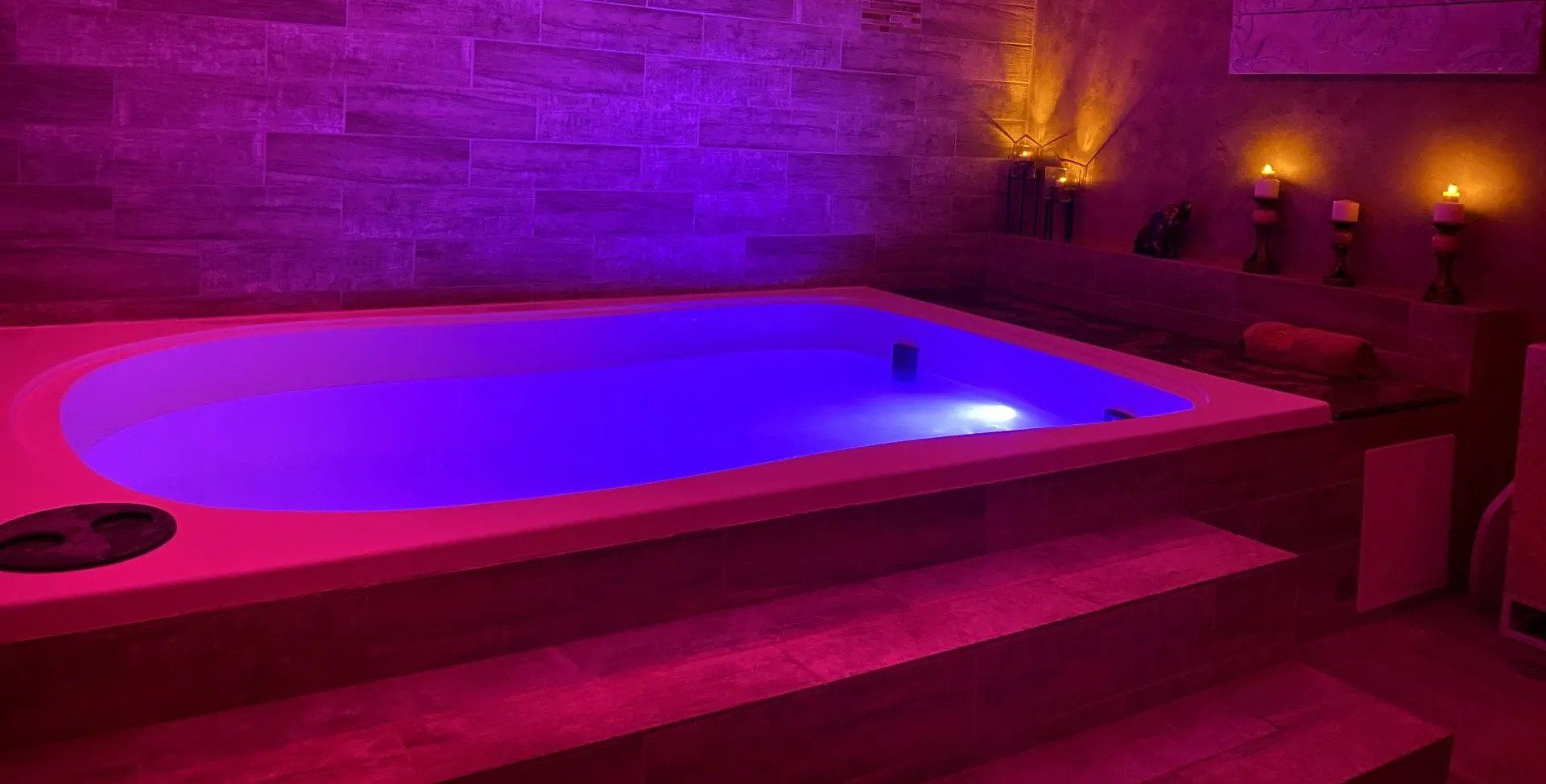 Indoor jacuzzi lit with purple and pink lights in a spa-like room with candles on the wall