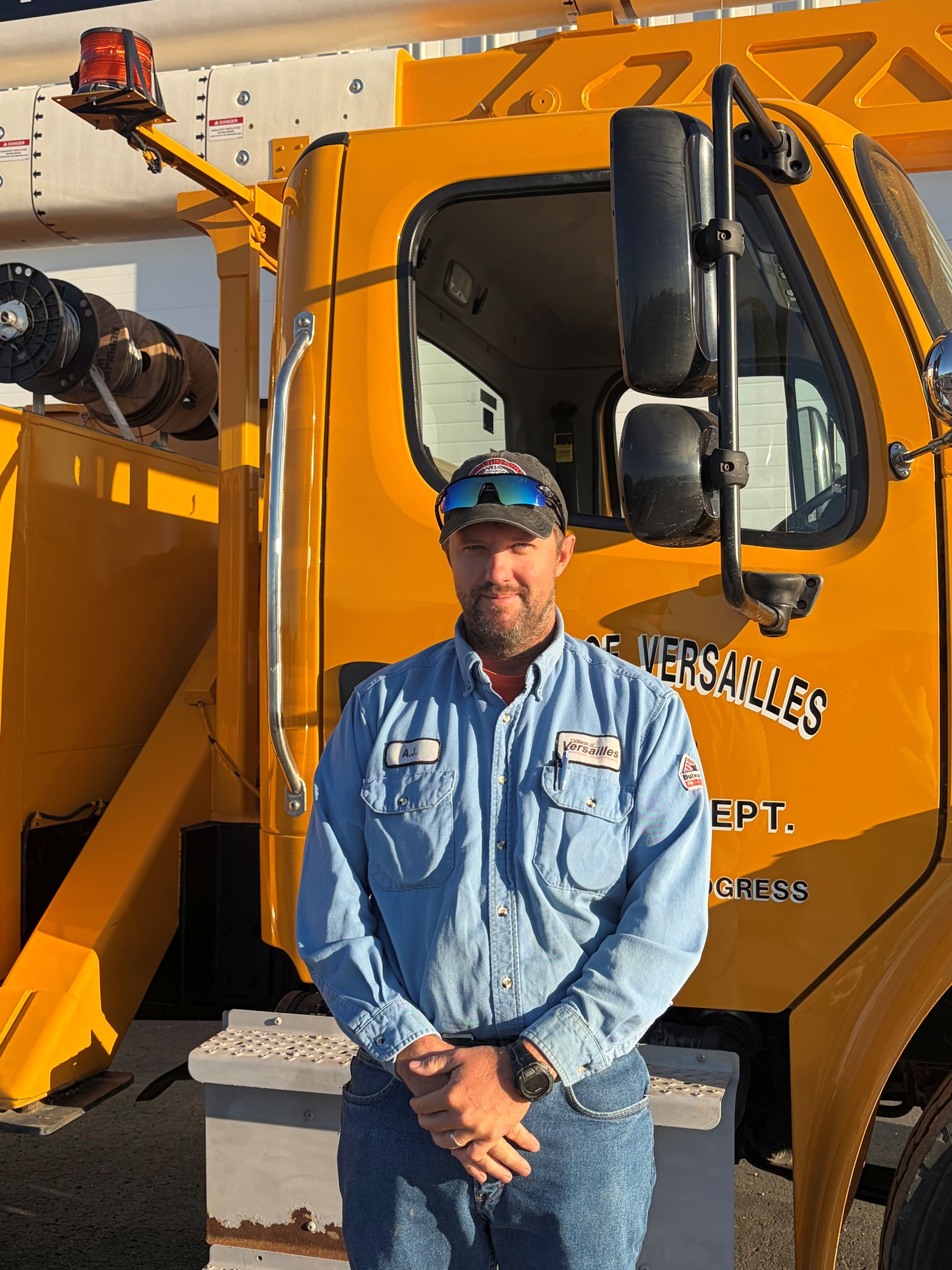 Smiling man in blue work shirt and cap with sunglasses, standing against a gray backdrop