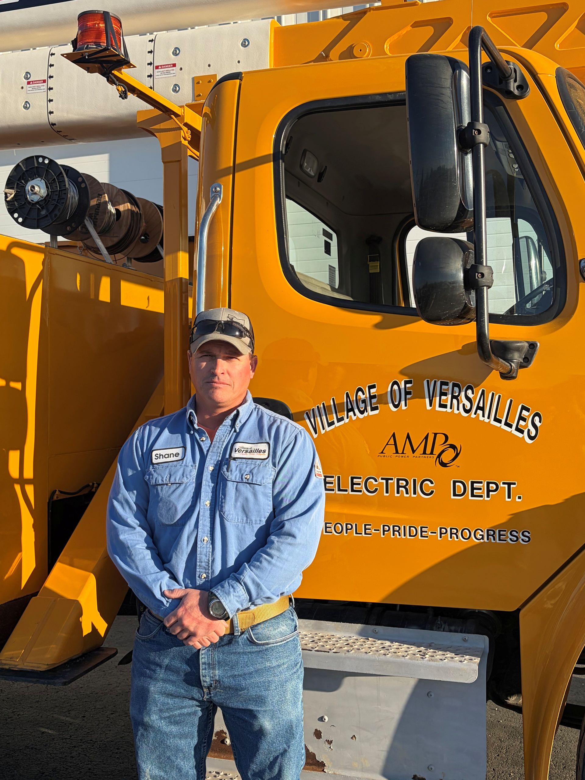 Man in blue work shirt and cap standing with hands clasped against a plain beige wall.