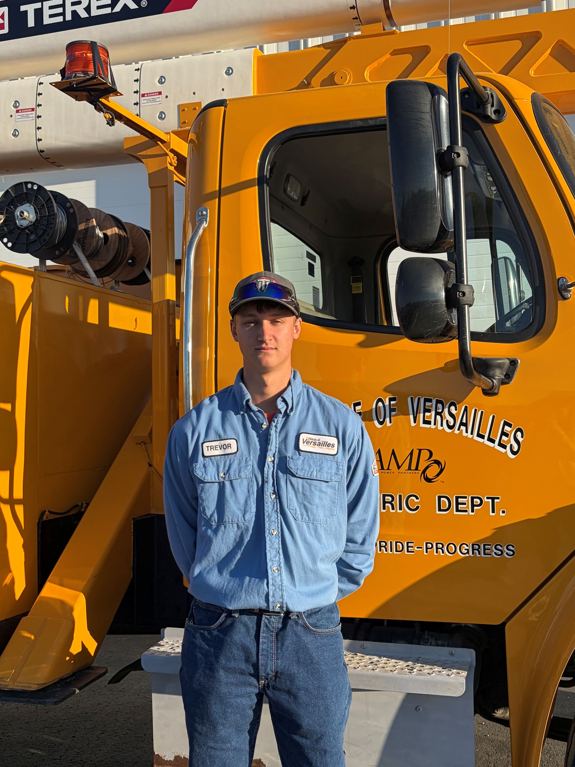 Man standing against a plain wall, wearing a blue work shirt, jeans, and a cap.