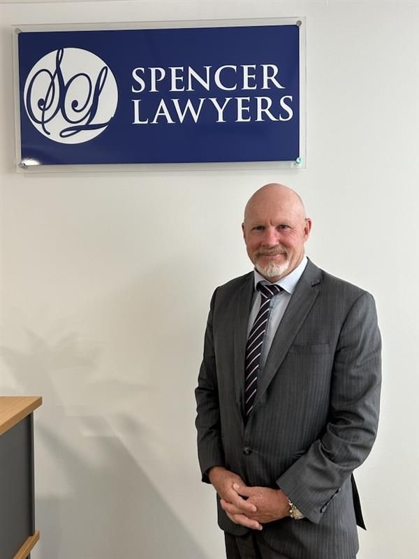 Man in a gray suit standing beside a Spencer Lawyers sign in an office hallway — Spencer Lawyers In Gosford, NSW