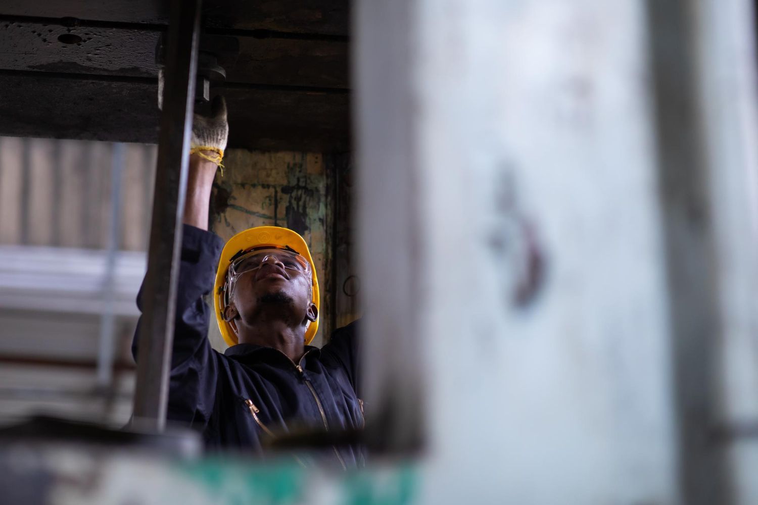 Worker in a yellow hard hat and safety glasses inspecting overhead in an industrial setting.