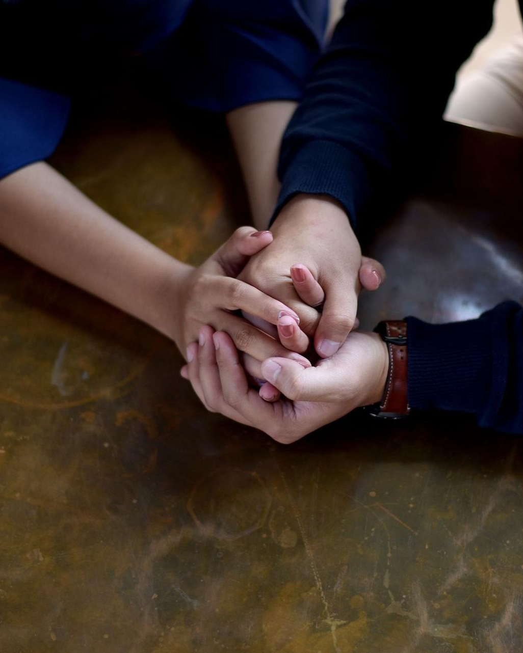 Hands of two people, one on top of the other, intertwined on a brown surface.