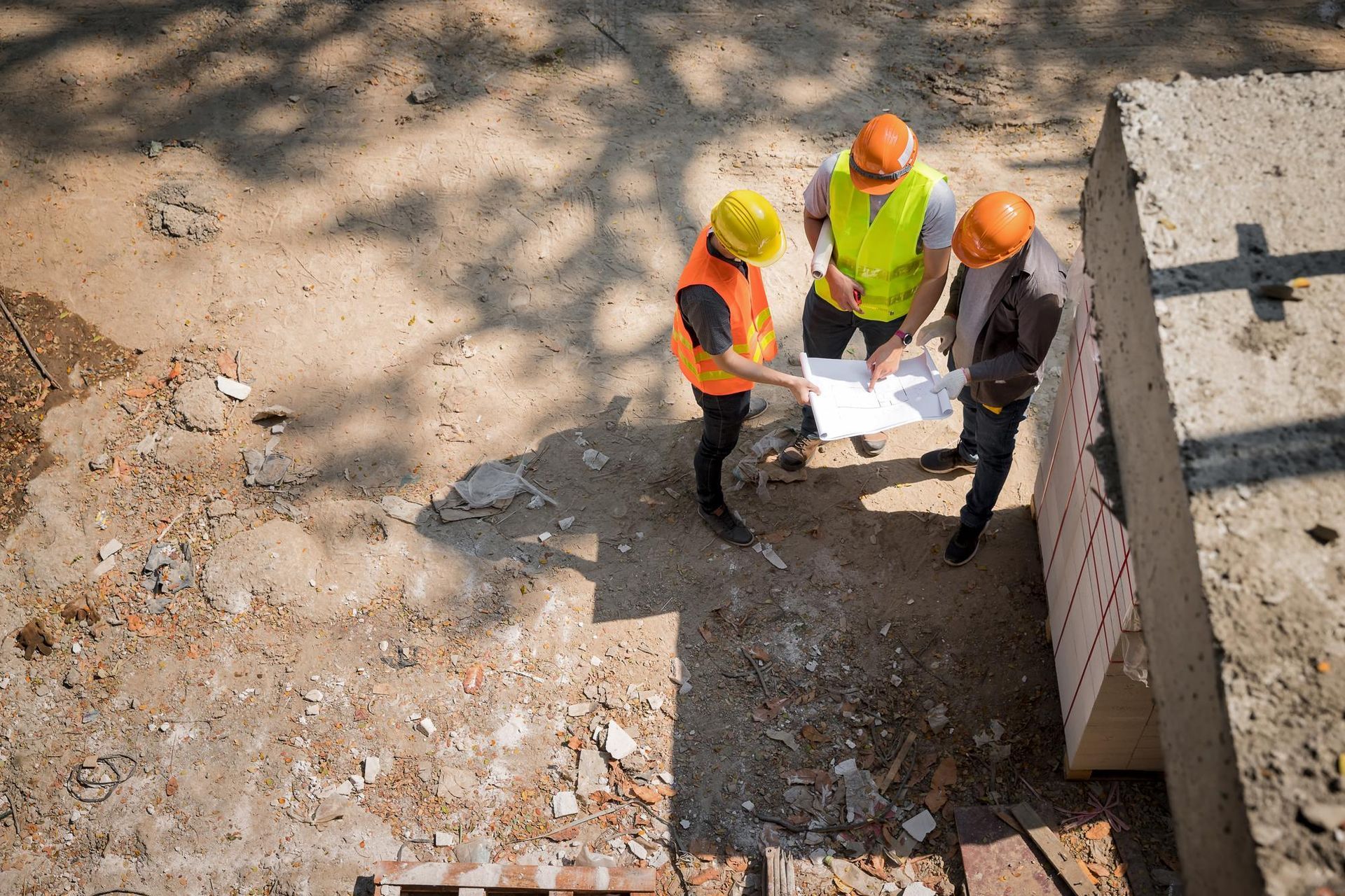 Three construction workers in hard hats and vests review blueprints on a construction site.