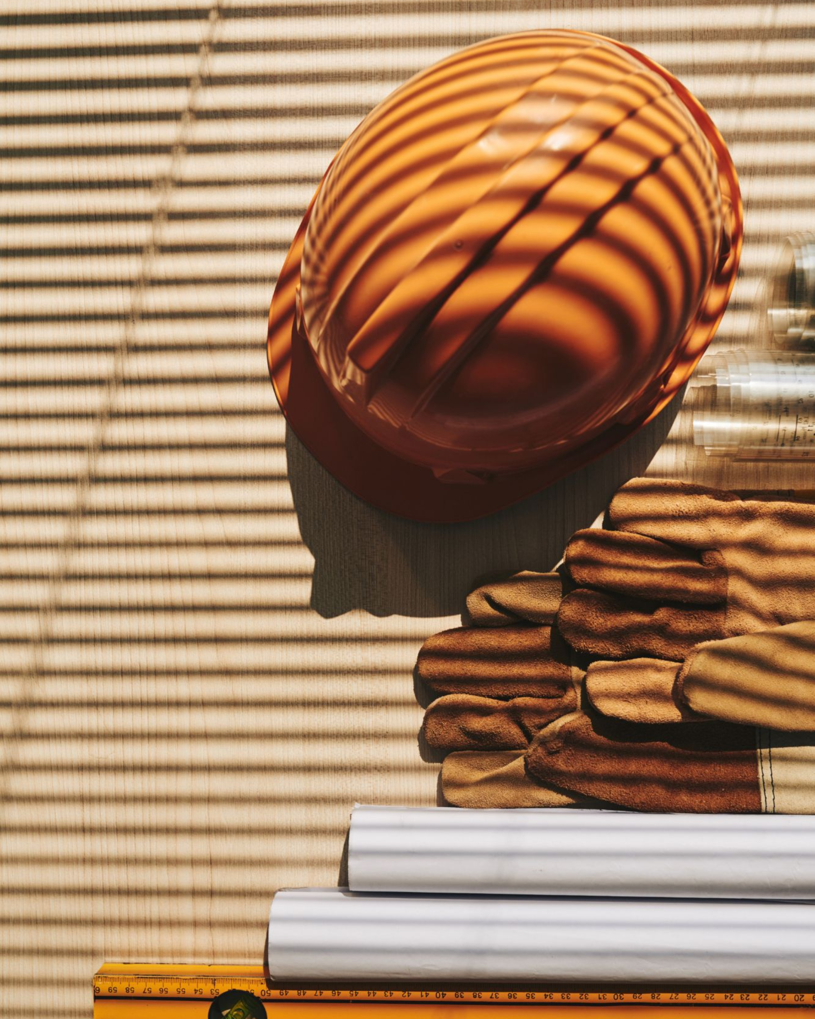 Orange hard hat, gloves, and blueprints on a construction site, with shadow from blinds.