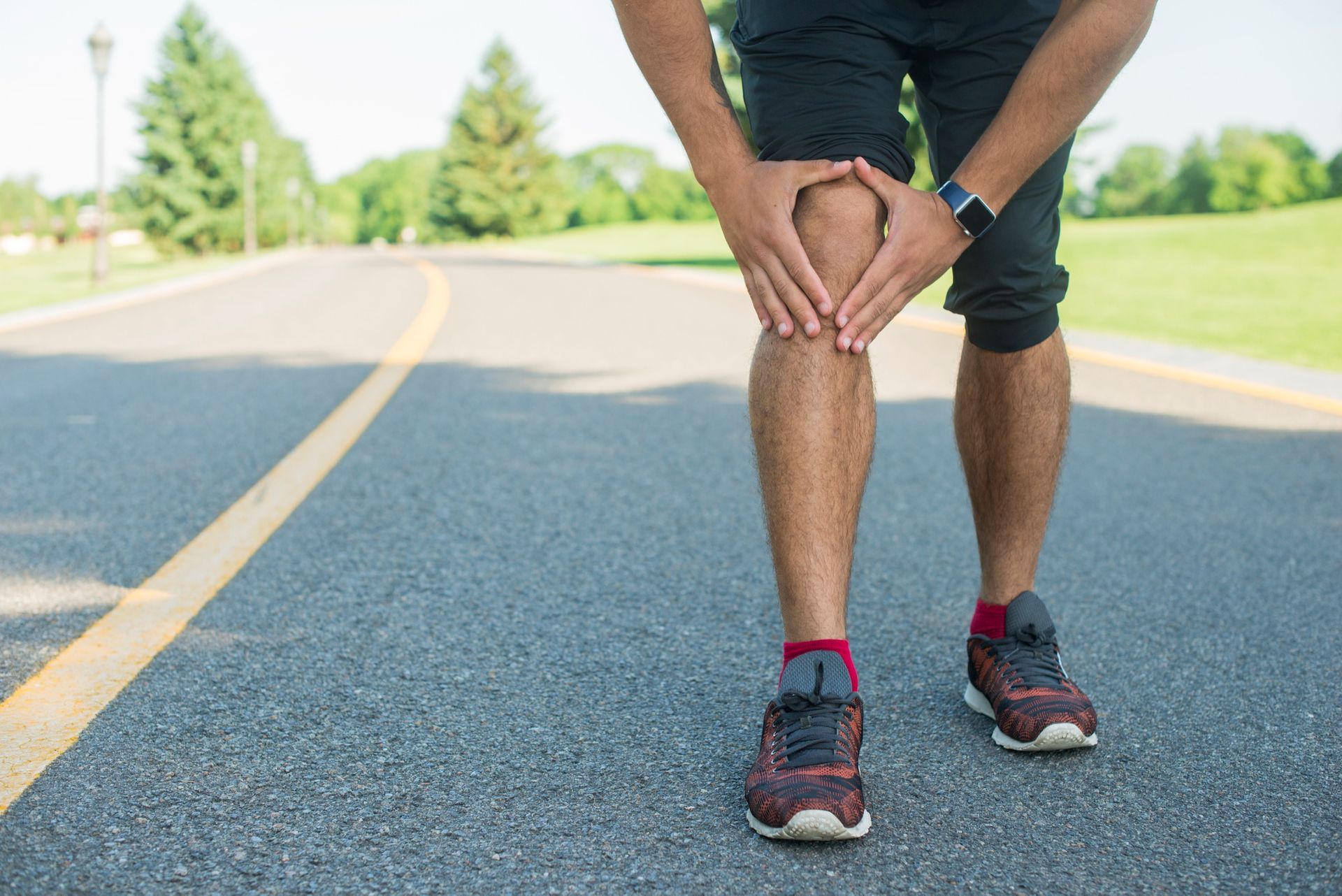 Person gripping both knees, possibly experiencing knee pain, on a paved path in a park.