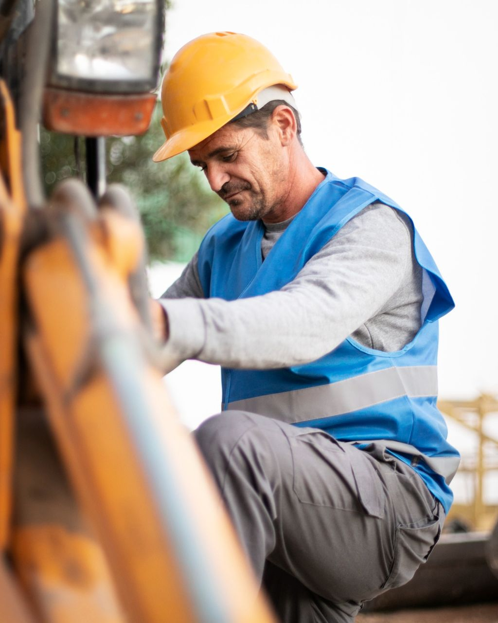 Construction worker in a yellow hard hat and blue vest leans over machinery, inspecting it outdoors.