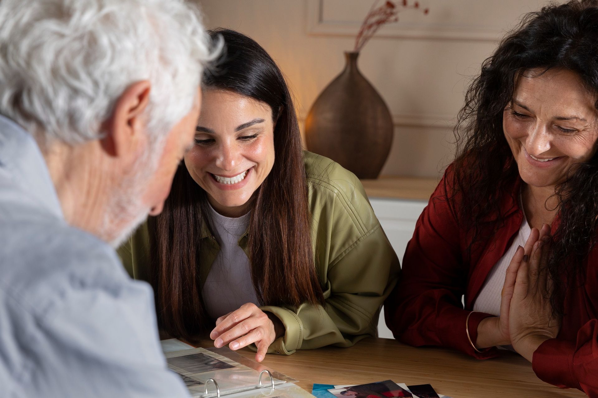 Three people looking at photos; one smiles, one has hands clasped, at a table.