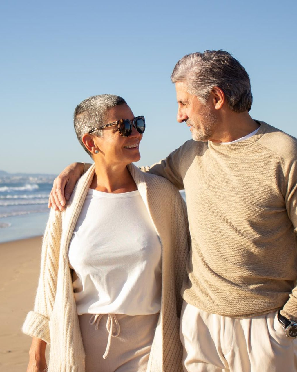 Couple walking on beach, man with arm around woman, looking at each other, smiling.