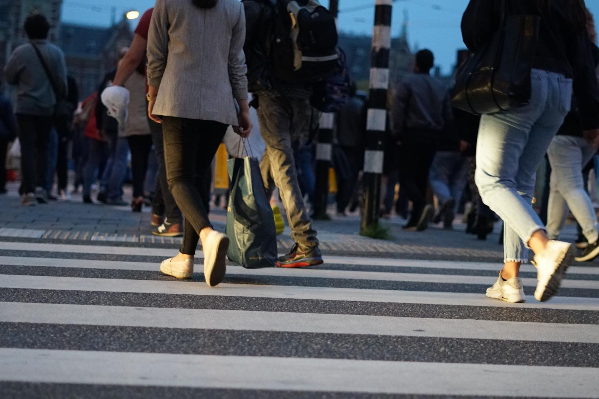 People walking on a zebra-striped crosswalk at dusk. Some wear backpacks, sneakers.