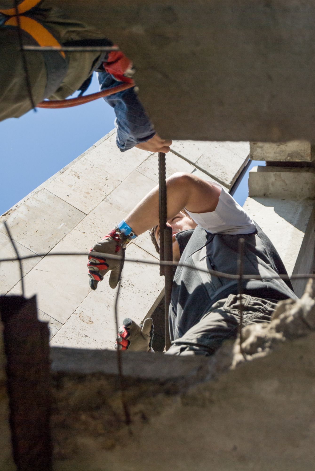 Construction worker reaching into opening, holding rebar, wearing gloves and safety harness.