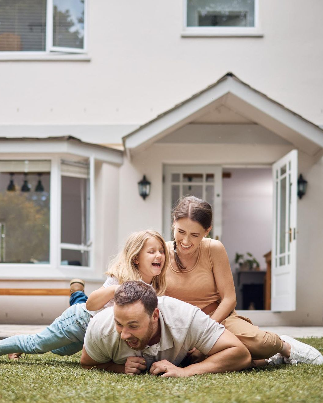 Family playing on grass in front of a house. Father on ground, child and mother smiling.