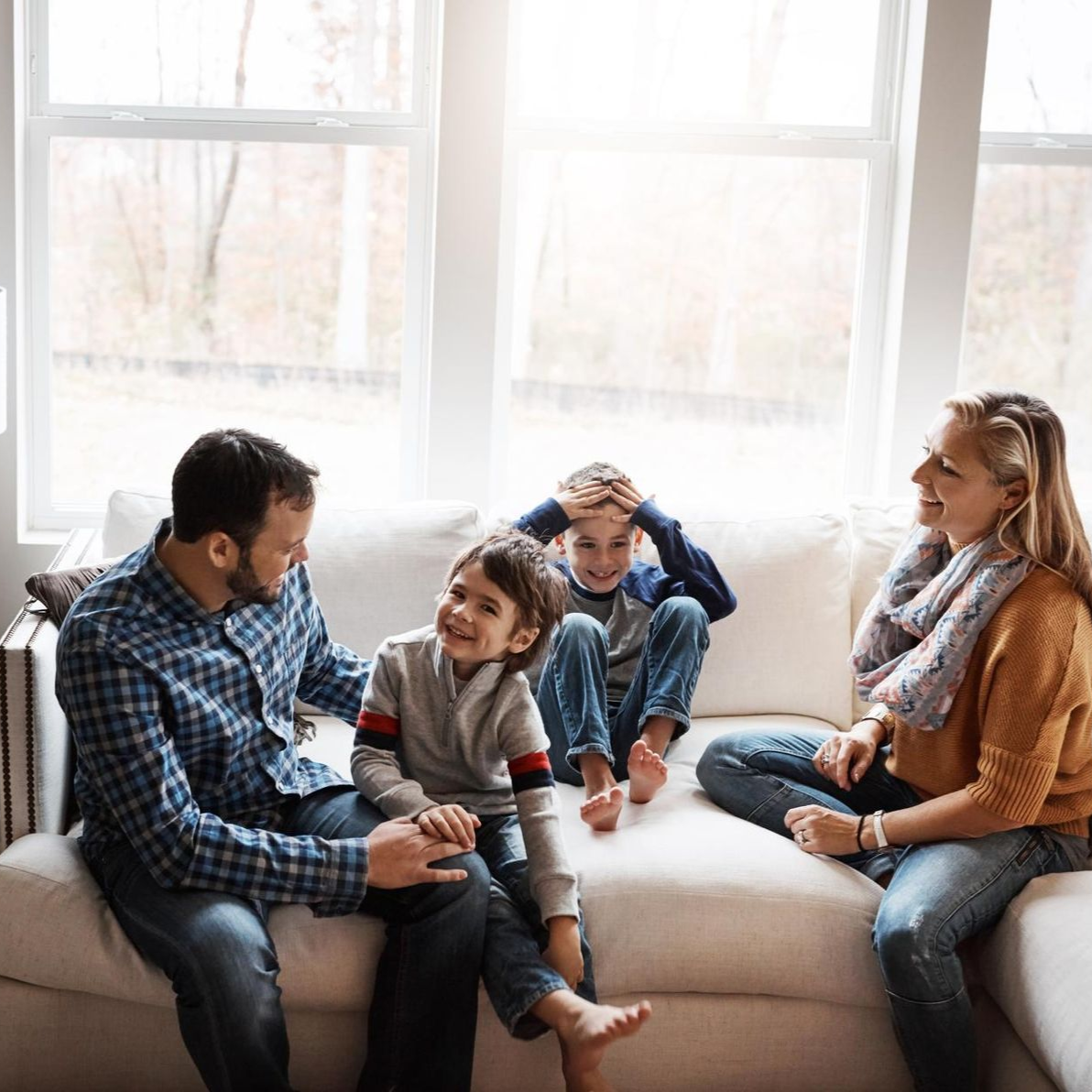Family of four sitting on a white sofa in a bright living room, smiling and interacting.