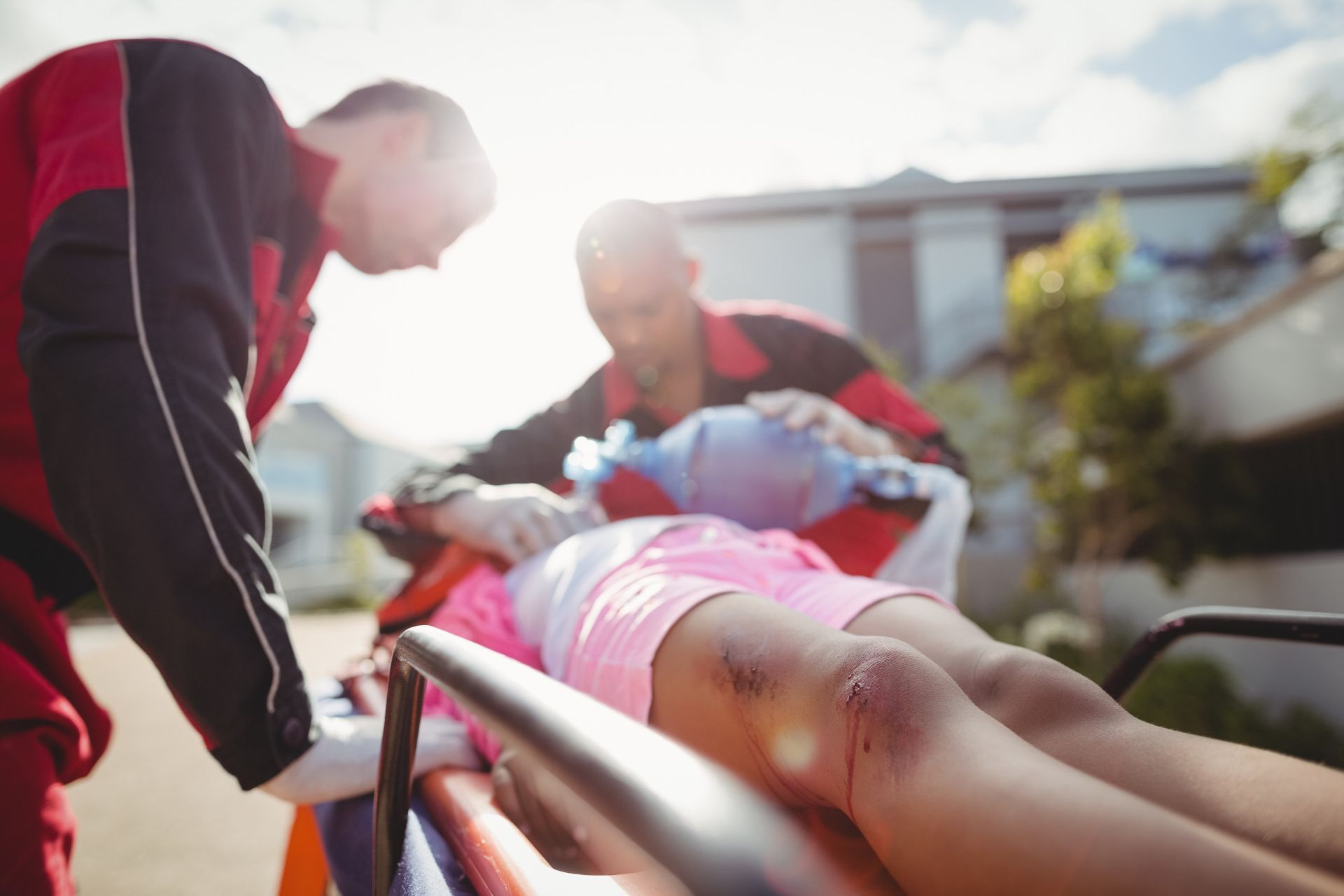 Paramedics tending to a patient on a stretcher outdoors. One holds a mask, other checks patient.
