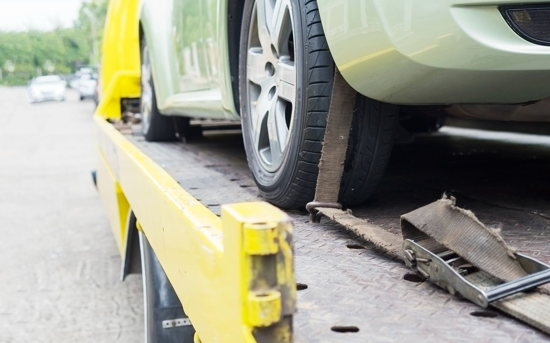 Green car on a yellow tow truck bed, secured with straps; daytime.