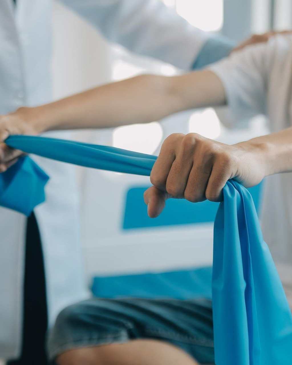 Therapist assisting a patient with arm exercises using a blue resistance band in a clinical setting.