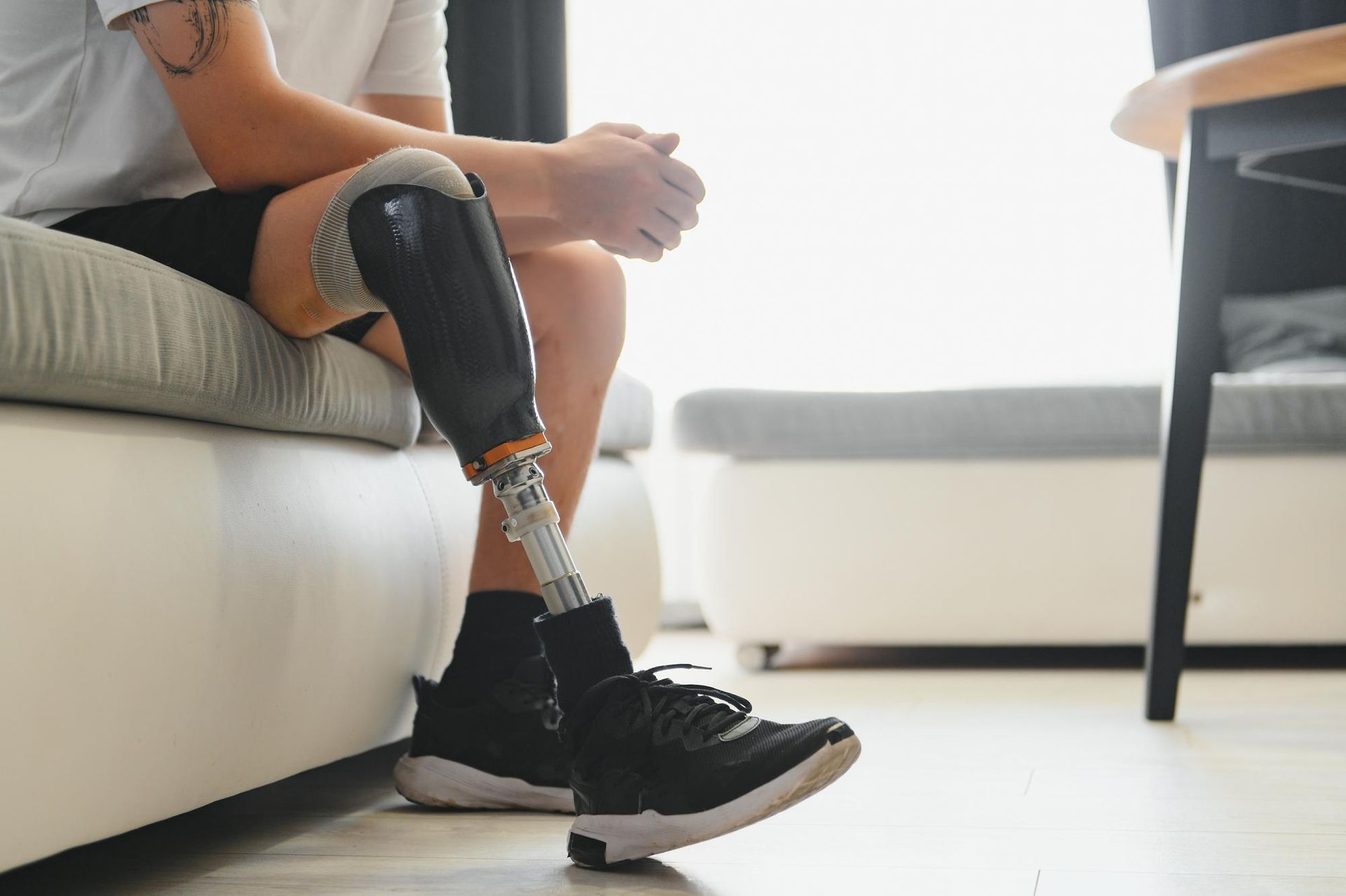 Person with prosthetic leg sits on a couch, hands clasped, wearing a black shoe.