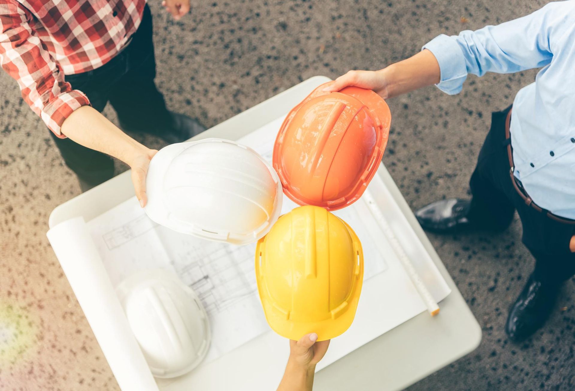 Three people holding hard hats of white, orange, and yellow over blueprints on a table.