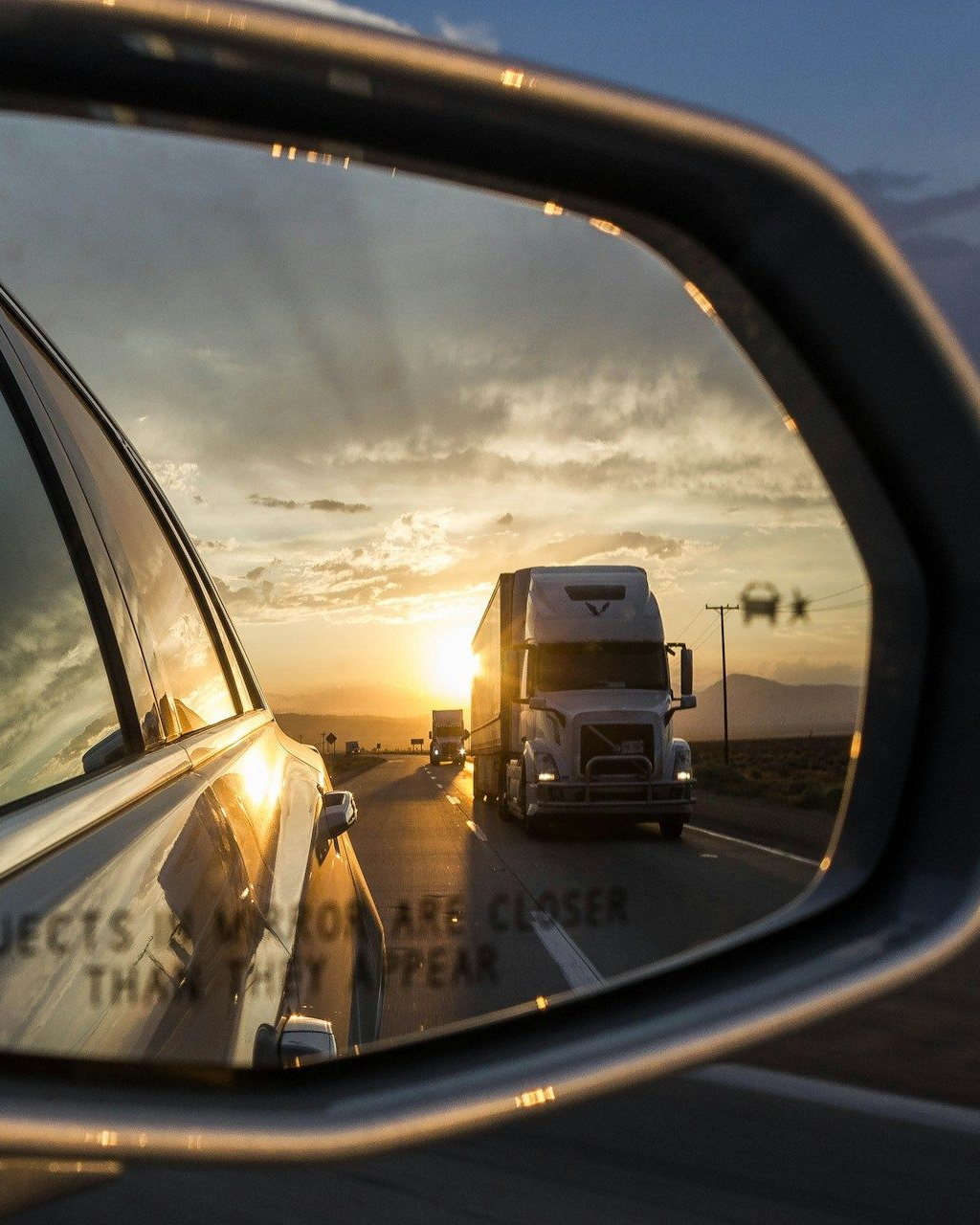 Side mirror view of highway with semi-truck driving toward the setting sun.