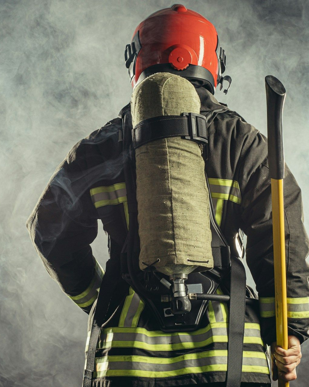 Firefighter in full gear, holding an axe, facing away from the camera, with smoky background.