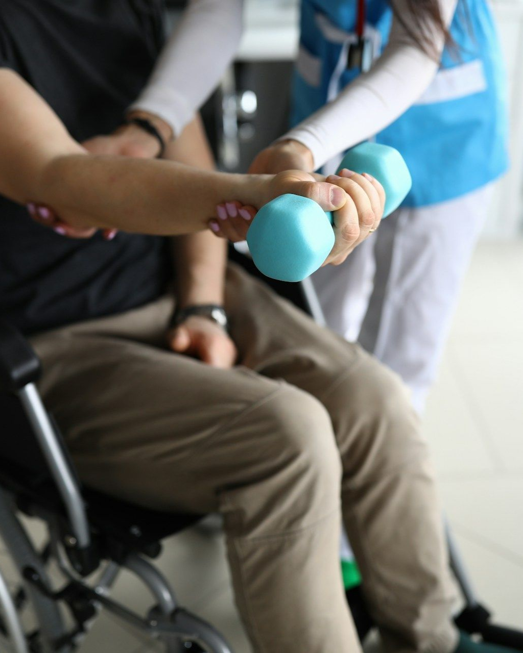 Therapist assists a person in a wheelchair lifting a small blue dumbbell for arm exercise.