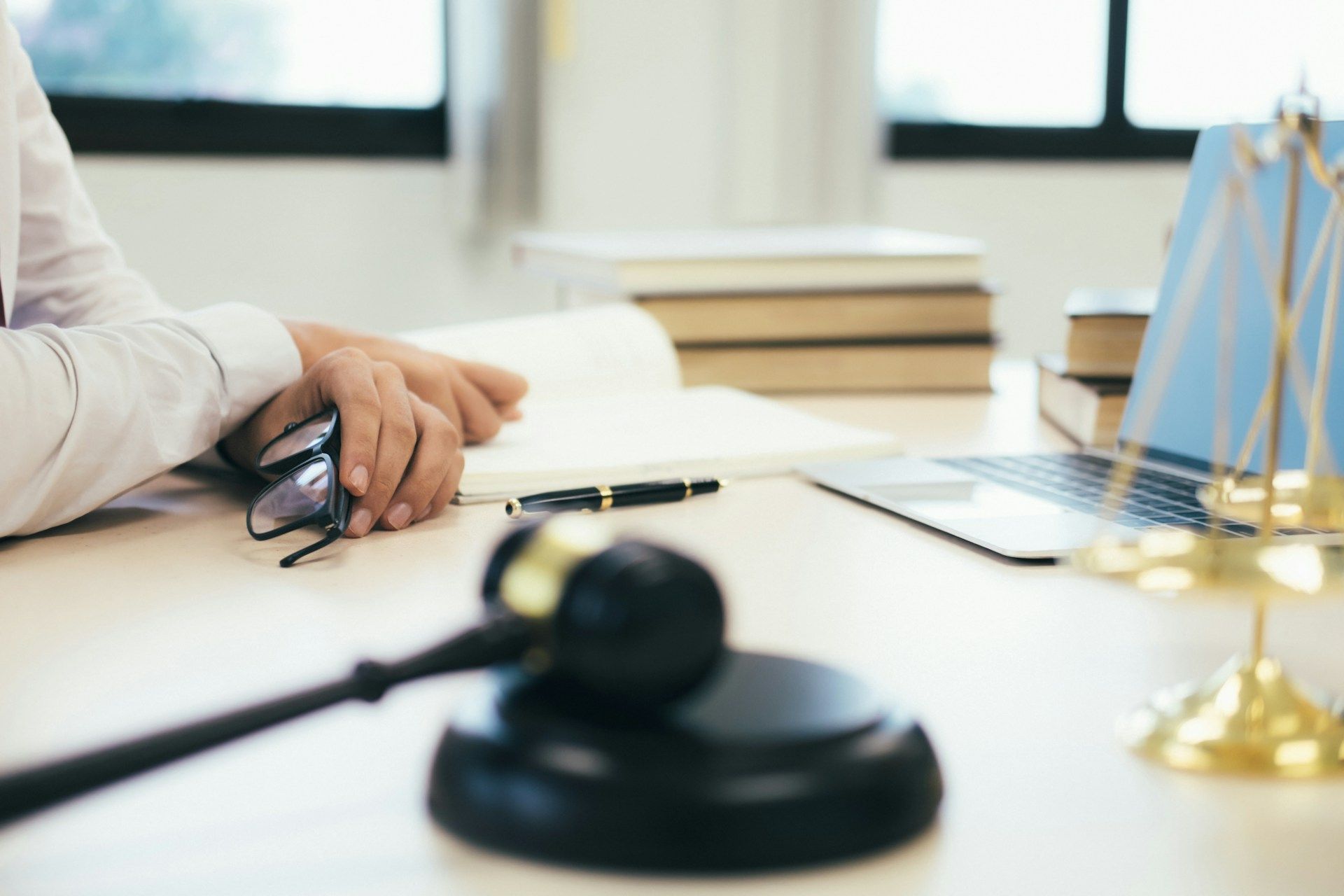 Gavel, scales of justice, laptop, and books on a desk, next to a person holding glasses and a document.