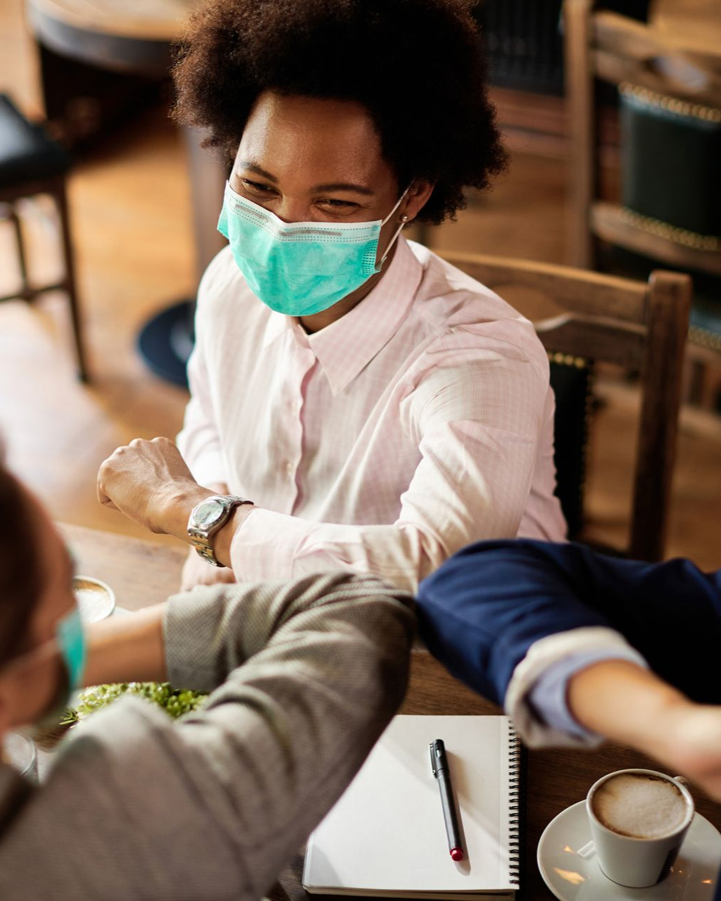 People in masks elbow bump at a table in a cafe, one person smiles.