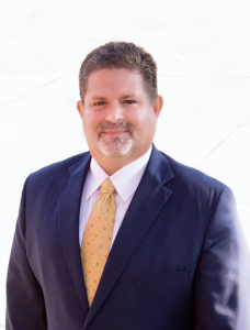 Man in blue suit, gold tie, smiling; outdoors, neutral background.